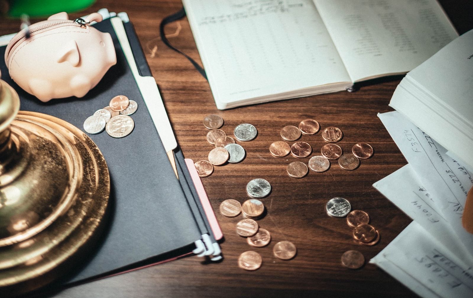 A ceramic piggy bank, a ledger notebook, and a small pile of coins spread across a wooden desk.