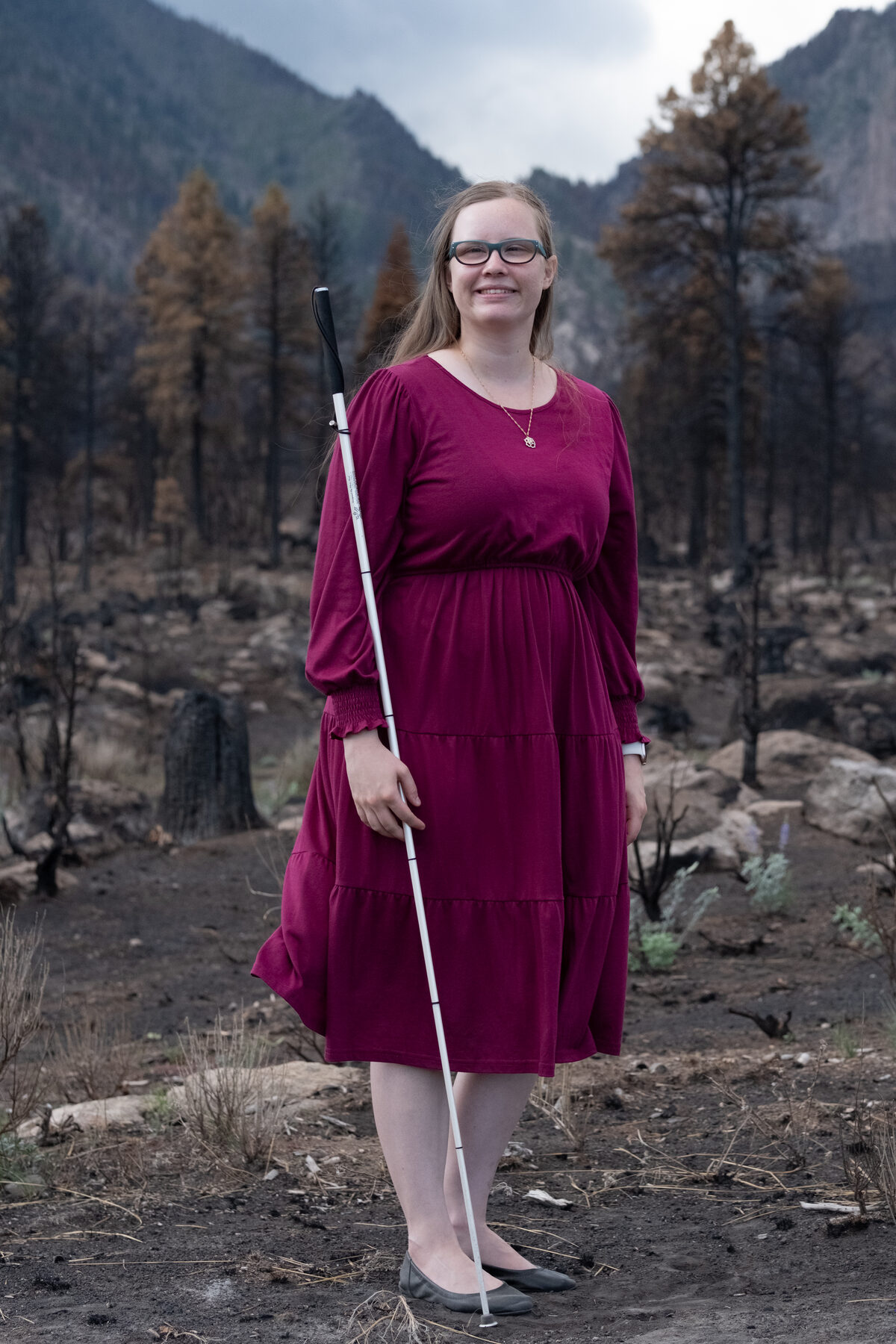 Brooke smiling outdoors in a warm maroon dress, holding her white cane, with a moody mountain forest behind her.